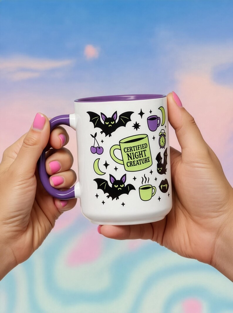 White ceramic mug with vibrant purple interior and matching C-handle. Features a full wrap-around, 'spooky cute' pastel goth design. Central graphic is a lime green coffee cup with 'CERTIFIED NIGHT CREATURE' in black text. Surrounding illustrations include grumpy black bats, a steaming green coffee cup, a purple mug, vampire fangs, crescent moon, black cherries, and an alarm clock. Design uses bold graphics in black, lime green, and purple, accented with small black stars. Pastel Goth vibes.