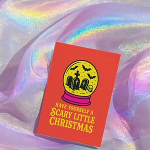 A vibrant, flat-lay photograph of the "Scary Little Christmas" greeting card. The card has a bright orange background with bold, retro yellow text. The central graphic is a snow globe with a yellow glass dome and a hot pink base. Inside the globe is a black silhouette of a spooky graveyard scene, featuring tombstones with crosses, a skull, and a swarm of flying bats. The card is made from premium 120lb silk cover stock with a slight sheen and measures 5x7 inches.