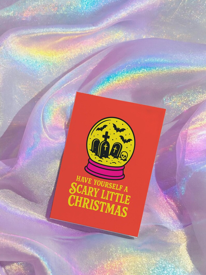 A vibrant, flat-lay photograph of the "Scary Little Christmas" greeting card. The card has a bright orange background with bold, retro yellow text. The central graphic is a snow globe with a yellow glass dome and a hot pink base. Inside the globe is a black silhouette of a spooky graveyard scene, featuring tombstones with crosses, a skull, and a swarm of flying bats. The card is made from premium 120lb silk cover stock with a slight sheen and measures 5x7 inches.