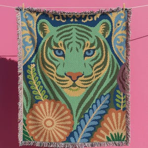 May include: A woven throw blanket featuring a tiger face design. The tiger is depicted in shades of green, blue, and yellow, with floral and leaf accents. The blanket has a fringed edge and is hanging against a pink background.