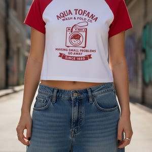 A women's fitted, mid-length micro rib raglan baby tee. The tee's body is solid white, made from a 1x1 micro-rib fabric.  It features short, set-in sleeves and a crewneck collar binding in a bold, contrasting athletic red. Centered on the chest is a vintage-inspired graphic printed in the same red ink. The design is for the "Aqua Tofana Wash & Fold Co." and features an illustration of an old-fashioned washing machine. Text below the machine reads "Making small problems go away" and "Since 1680."