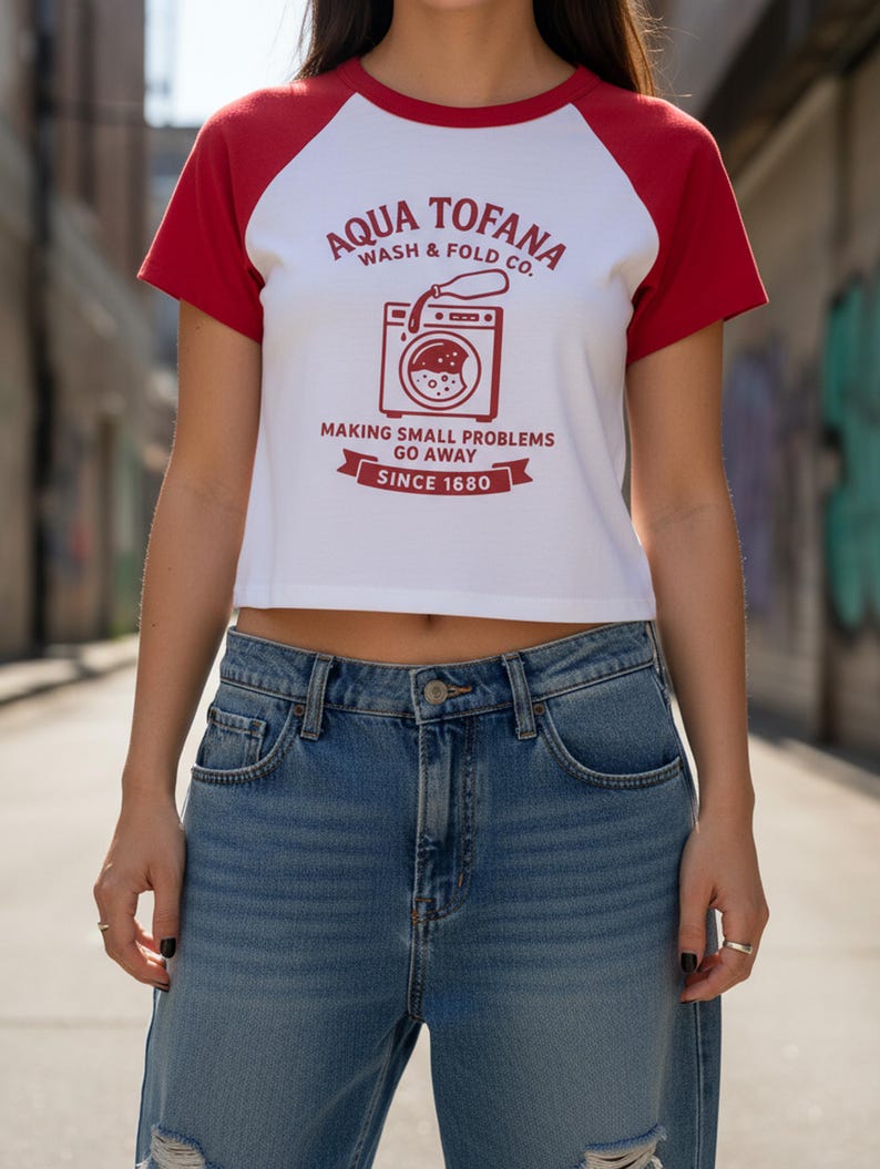 A women's fitted, mid-length micro rib raglan baby tee. The tee's body is solid white, made from a 1x1 micro-rib fabric.  It features short, set-in sleeves and a crewneck collar binding in a bold, contrasting athletic red. Centered on the chest is a vintage-inspired graphic printed in the same red ink. The design is for the "Aqua Tofana Wash & Fold Co." and features an illustration of an old-fashioned washing machine. Text below the machine reads "Making small problems go away" and "Since 1680."