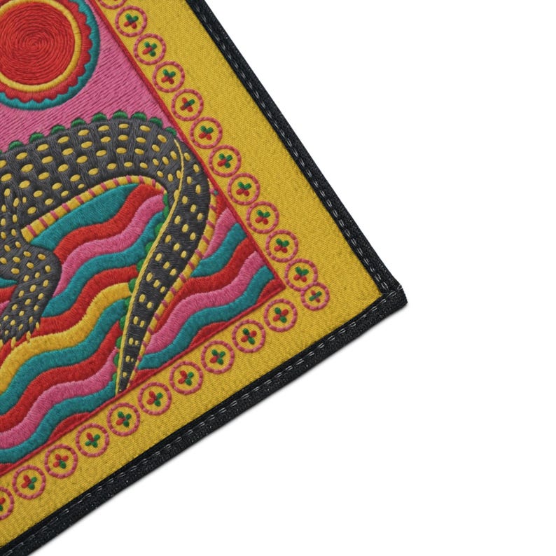 A rectangular doormat featuring a colorful 2D flat print designed to mimic raised, textured yarn embroidery. The central graphic showcases a large dark grey and yellow alligator resting in wavy water stripes of red, bright blue, and yellow. The background is hot pink, decorated with two green palm trees, a leafy green agave plant, and two stylized suns. The entire design is enclosed by a yellow border with repeating circle motifs and finished with a sleek black outer edge border trim.