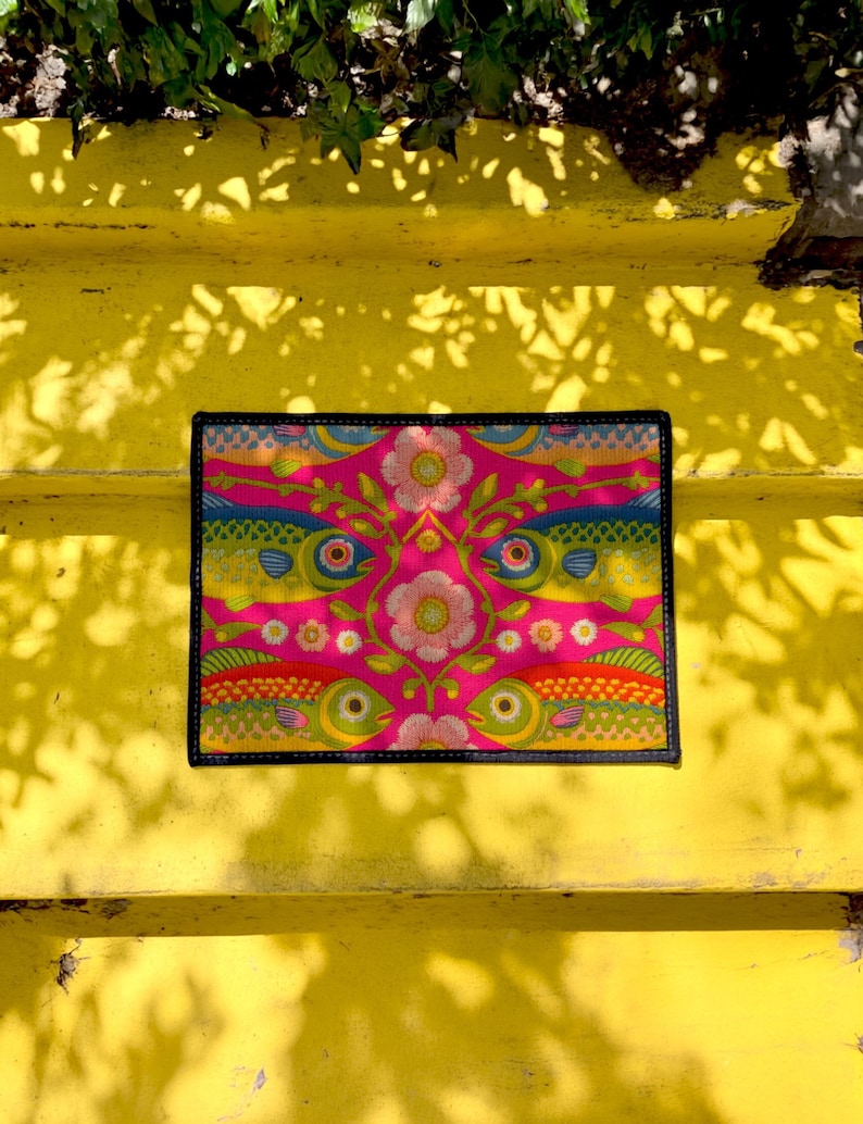 Rectangular indoor outdoor doormat featuring a vibrant hot pink background with a symmetrical maximalist pattern. The design showcases stylized sardines in yellow, lime green, and cobalt blue swimming horizontally towards a central vertical axis of blooming pale pink flowers and green vines. The imagery mimics the texture of vintage needlepoint embroidery but is a flat print on polyester. The mat is framed by a sleek black trim and features a non-slip rubber backing for safety.