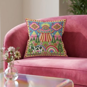 Description of a square throw pillow with a vibrant maximalist alien design. A central UFO with a large all-seeing eye hovers above a landscape with two rainbows, pink mountains, and green pine trees. A black and white spotted cow stands in the foreground. The design is a high-contrast palette of sky blue, magenta, hot pink, and meadow green. The pillow has a faux-embroidery 2D print finish on linen-feel fabric, framed by a yellow and pink scalloped floral border.