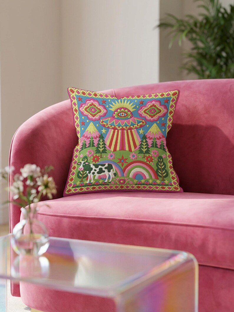 Description of a square throw pillow with a vibrant maximalist alien design. A central UFO with a large all-seeing eye hovers above a landscape with two rainbows, pink mountains, and green pine trees. A black and white spotted cow stands in the foreground. The design is a high-contrast palette of sky blue, magenta, hot pink, and meadow green. The pillow has a faux-embroidery 2D print finish on linen-feel fabric, framed by a yellow and pink scalloped floral border.