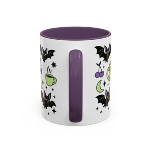 White ceramic mug with vibrant purple interior and matching C-handle. Features a full wrap-around, 'spooky cute' pastel goth design. Central graphic is a lime green coffee cup with 'CERTIFIED NIGHT CREATURE' in black text. Surrounding illustrations include grumpy black bats, a steaming green coffee cup, a purple mug, vampire fangs, crescent moon, black cherries, and an alarm clock. Design uses bold graphics in black, lime green, and purple, accented with small black stars. Pastel Goth vibes.