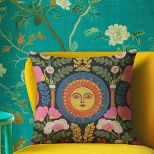A square folk art sun throw pillow with a dark, charcoal-gray background. The central design is a vibrant celestial sun face in shades of orange, yellow, and red, with defined facial features. The sun is enclosed in a circular blue border with a textured, faux-embroidery pattern. Surrounding the central sun are symmetrical, colorful botanical and floral elements, including pink and white flowers, and green and gold leaves. The overall style is maximalist and bohemian.