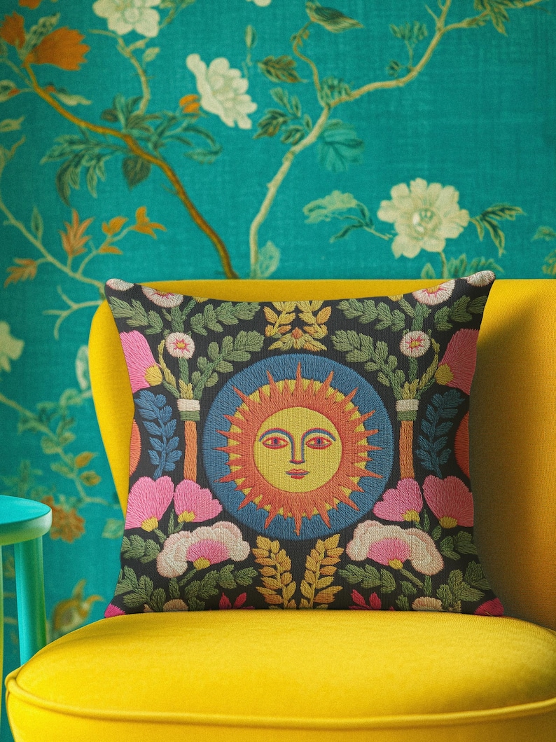 A square folk art sun throw pillow with a dark, charcoal-gray background. The central design is a vibrant celestial sun face in shades of orange, yellow, and red, with defined facial features. The sun is enclosed in a circular blue border with a textured, faux-embroidery pattern. Surrounding the central sun are symmetrical, colorful botanical and floral elements, including pink and white flowers, and green and gold leaves. The overall style is maximalist and bohemian.