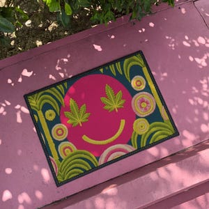 A rectangular doormat featuring a large hot pink smiley face on a deep teal background. The smiley face has green cannabis leaves for eyes and a yellow stitched-style smile. The central face is surrounded by stylized green foliage arches and concentric circles in pink, yellow, and white, resembling abstract flowers. The design mimics a vintage needlepoint or tapestry texture but is a flat print. The mat has a solid black trim border.