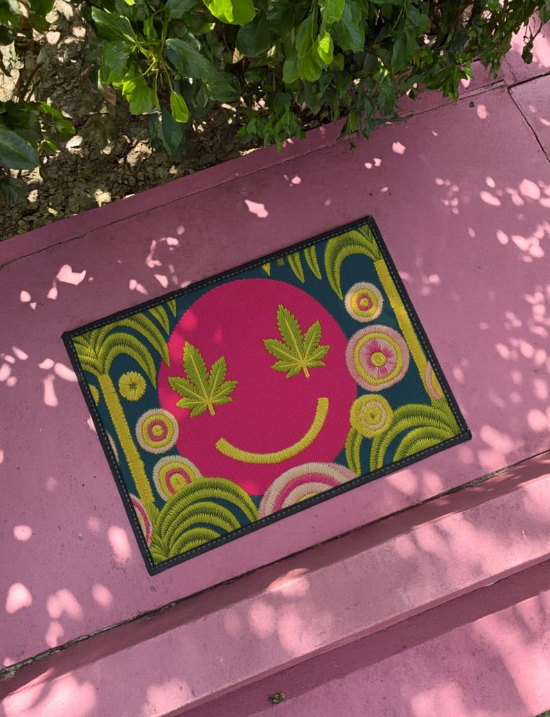 A rectangular doormat featuring a large hot pink smiley face on a deep teal background. The smiley face has green cannabis leaves for eyes and a yellow stitched-style smile. The central face is surrounded by stylized green foliage arches and concentric circles in pink, yellow, and white, resembling abstract flowers. The design mimics a vintage needlepoint or tapestry texture but is a flat print. The mat has a solid black trim border.