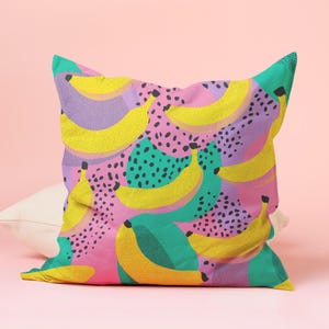 Puede incluir: Un coj&iacute;n cuadrado decorativo con un vibrante estampado de pl&aacute;tanos. El dise&ntilde;o presenta pl&aacute;tanos amarillos sobre un fondo de formas rosas, turquesas y moradas, acentuado con lunares negros. Un elemento de decoraci&oacute;n del hogar brillante y colorido.