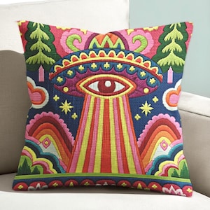 Puede incluir: Cojín bordado con un diseño colorido de OVNI con un ojo en el centro, irradiando sobre un paisaje de arcoíris. El cojín tiene un fondo azul marino con detalles en rosa, amarillo, verde y rojo. El cojín es cuadrado.
