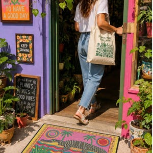 A rectangular doormat featuring a colorful 2D flat print designed to mimic raised, textured yarn embroidery. The central graphic showcases a large dark grey and yellow alligator resting in wavy water stripes of red, bright blue, and yellow. The background is hot pink, decorated with two green palm trees, a leafy green agave plant, and two stylized suns. The entire design is enclosed by a yellow border with repeating circle motifs and finished with a sleek black outer edge border trim.