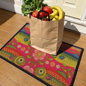 Rectangular indoor outdoor doormat featuring a vibrant hot pink background with a symmetrical maximalist pattern. The design showcases stylized sardines in yellow, lime green, and cobalt blue swimming horizontally towards a central vertical axis of blooming pale pink flowers and green vines. The imagery mimics the texture of vintage needlepoint embroidery but is a flat print on polyester. The mat is framed by a sleek black trim and features a non-slip rubber backing for safety.