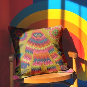 A square faux embroidery pillow featuring a brightly colored maximalist design against a mustard yellow background. In the center stands a stylized, furry Bigfoot or Sasquatch figure rendered in vibrant layers of pink, orange, blue, and lime green with a blue face and yellow eyes. The central creature is surrounded by large, detailed symmetrical pink and blue flowers and leaves. The flat printed fabric creates a highly realistic optical illusion of dimensional crewel embroidery stitches.