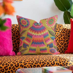 A square faux embroidery pillow featuring a brightly colored maximalist design against a mustard yellow background. In the center stands a stylized, furry Bigfoot or Sasquatch figure rendered in vibrant layers of pink, orange, blue, and lime green with a blue face and yellow eyes. The central creature is surrounded by large, detailed symmetrical pink and blue flowers and leaves. The flat printed fabric creates a highly realistic optical illusion of dimensional crewel embroidery stitches.