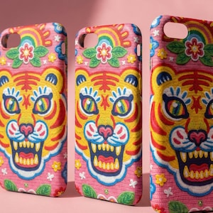 May include: Three pink phone cases with a colorful tiger face design. The cases feature a yellow tiger face with blue, red, and white accents, a rainbow, and floral details. The cases are designed for a smartphone.