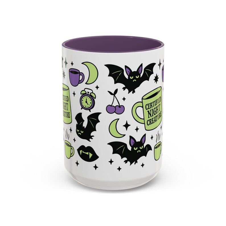 White ceramic mug with vibrant purple interior and matching C-handle. Features a full wrap-around, 'spooky cute' pastel goth design. Central graphic is a lime green coffee cup with 'CERTIFIED NIGHT CREATURE' in black text. Surrounding illustrations include grumpy black bats, a steaming green coffee cup, a purple mug, vampire fangs, crescent moon, black cherries, and an alarm clock. Design uses bold graphics in black, lime green, and purple, accented with small black stars. Pastel Goth vibes.