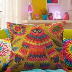 A square faux embroidery pillow featuring a brightly colored maximalist design against a mustard yellow background. In the center stands a stylized, furry Bigfoot or Sasquatch figure rendered in vibrant layers of pink, orange, blue, and lime green with a blue face and yellow eyes. The central creature is surrounded by large, detailed symmetrical pink and blue flowers and leaves. The flat printed fabric creates a highly realistic optical illusion of dimensional crewel embroidery stitches.