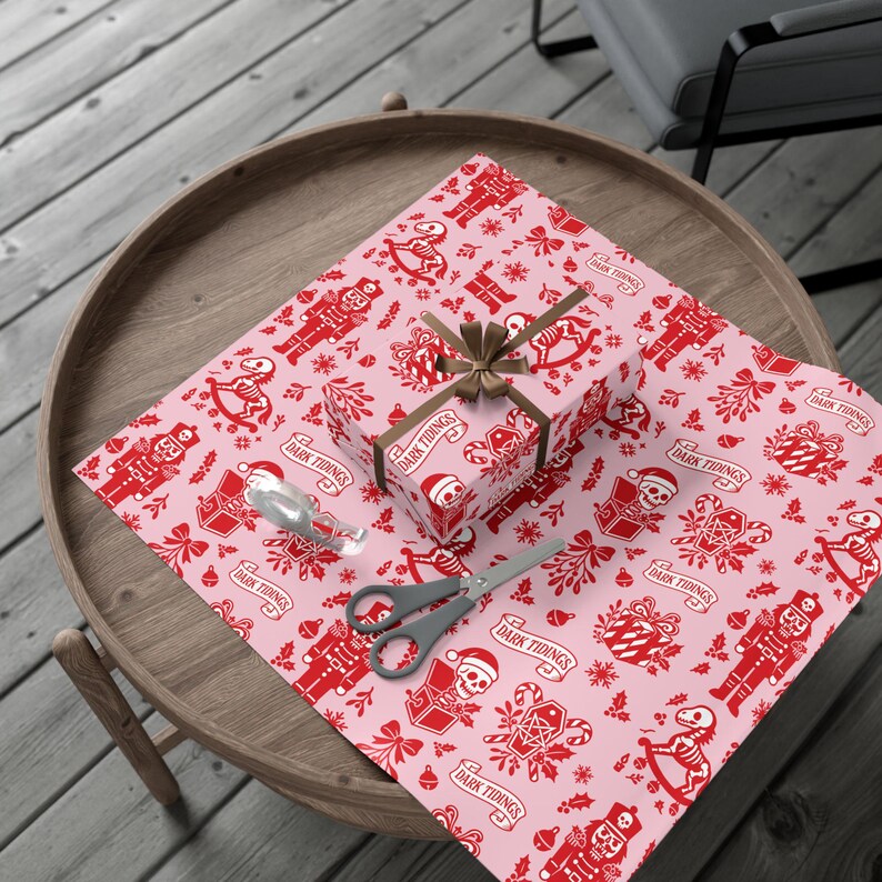 Premium satin finish wrapping paper featuring a repeating pastel goth pattern on a pink background. The design showcases a playful yet spooky aesthetic with high-contrast skeleton nutcrackers, skull jack-in-the-boxes, and rocking horse skeletons in soft pinks, and reds. The paper texture appears semi-glossy. Shown in three available sizes including sheets and rolls, specifically 29 inches wide, suitable for wrapping gifts for holidays, birthdays, or gothic themed occasions.