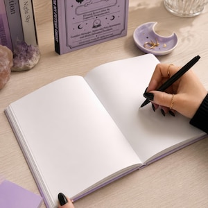 A 5 by 7 inch hardcover journal with a matte pastel purple cover and black printed graphics. The front features a retro inspired illustration of a white ghost character. One design shows the ghost holding a locked book flanked by a lit candle and a skeleton key with text reading Notes For My Future Ghost. The other shows the ghost peeking over a blank label with text reading Property Of The Future Ghost. Both include crescent moons, four point stars, and vintage decorative border frames.