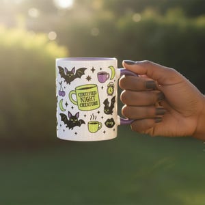 White ceramic mug with vibrant purple interior and matching C-handle. Features a full wrap-around, 'spooky cute' pastel goth design. Central graphic is a lime green coffee cup with 'CERTIFIED NIGHT CREATURE' in black text. Surrounding illustrations include grumpy black bats, a steaming green coffee cup, a purple mug, vampire fangs, crescent moon, black cherries, and an alarm clock. Design uses bold graphics in black, lime green, and purple, accented with small black stars. Pastel Goth vibes.