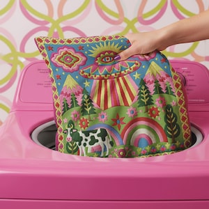 Description of a square throw pillow with a vibrant maximalist alien design. A central UFO with a large all-seeing eye hovers above a landscape with two rainbows, pink mountains, and green pine trees. A black and white spotted cow stands in the foreground. The design is a high-contrast palette of sky blue, magenta, hot pink, and meadow green. The pillow has a faux-embroidery 2D print finish on linen-feel fabric, framed by a yellow and pink scalloped floral border.