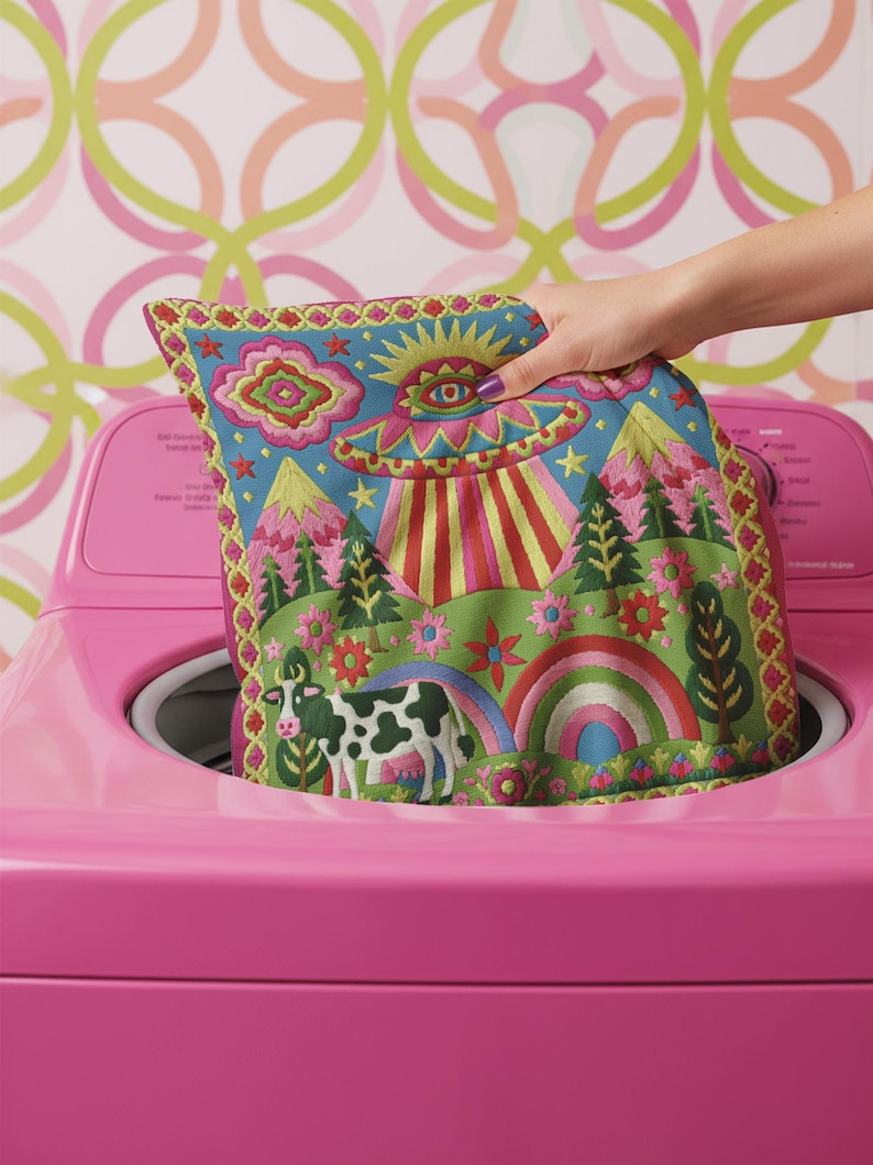 Description of a square throw pillow with a vibrant maximalist alien design. A central UFO with a large all-seeing eye hovers above a landscape with two rainbows, pink mountains, and green pine trees. A black and white spotted cow stands in the foreground. The design is a high-contrast palette of sky blue, magenta, hot pink, and meadow green. The pillow has a faux-embroidery 2D print finish on linen-feel fabric, framed by a yellow and pink scalloped floral border.