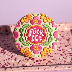 May include: Embroidered round patch with the text "FUCK ICE" in white, surrounded by a floral design in pink, yellow, and green. The patch is on a light beige background.
