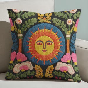 A square folk art sun throw pillow with a dark, charcoal-gray background. The central design is a vibrant celestial sun face in shades of orange, yellow, and red, with defined facial features. The sun is enclosed in a circular blue border with a textured, faux-embroidery pattern. Surrounding the central sun are symmetrical, colorful botanical and floral elements, including pink and white flowers, and green and gold leaves. The overall style is maximalist and bohemian.