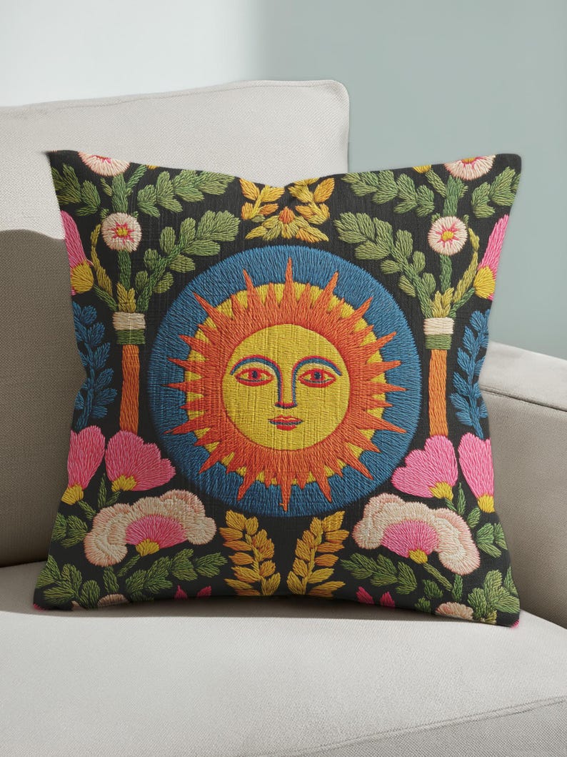 A square folk art sun throw pillow with a dark, charcoal-gray background. The central design is a vibrant celestial sun face in shades of orange, yellow, and red, with defined facial features. The sun is enclosed in a circular blue border with a textured, faux-embroidery pattern. Surrounding the central sun are symmetrical, colorful botanical and floral elements, including pink and white flowers, and green and gold leaves. The overall style is maximalist and bohemian.