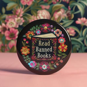 Może przedstawiać: Czarny guzik z kolorowym haftem przedstawiającym książkę z napisem "Read Banned Books" otoczoną kwiatami.