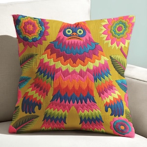 May include: Square cushion with a colourful embroidered design. The cushion features a vibrant, multi-coloured monster-like figure with floral and leaf accents on a mustard yellow background. The cushion is placed on a white sofa.