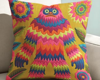 May include: Square decorative pillow with a colorful embroidered design. The pillow features a vibrant, multi-colored monster-like figure with floral and leaf accents on a mustard yellow background. The pillow is placed on a white sofa.
