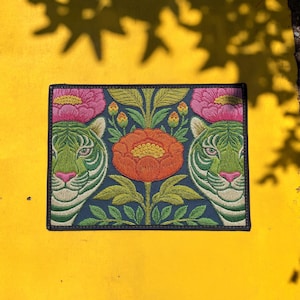 May include: Embroidered patch featuring two green tiger faces flanking a central orange flower. Pink flowers and green leaves are also present on a navy blue background. The patch is rectangular with a black border.