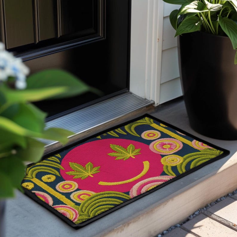 A rectangular doormat featuring a large hot pink smiley face on a deep teal background. The smiley face has green cannabis leaves for eyes and a yellow stitched-style smile. The central face is surrounded by stylized green foliage arches and concentric circles in pink, yellow, and white, resembling abstract flowers. The design mimics a vintage needlepoint or tapestry texture but is a flat print. The mat has a solid black trim border.