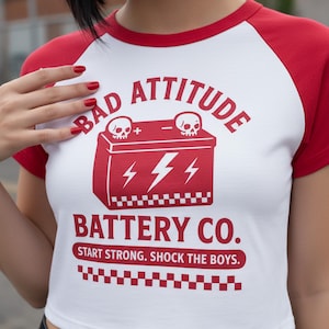 Könnte beinhalten: Weißes und rotes Raglan-Crop-Top mit dem roten Text "BAD ATTITUDE BATTERY CO.". Die Grafik zeigt eine Batterie mit Totenköpfen und Blitzen. Das Shirt hat unten ein Schachbrettmuster.