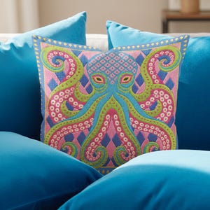 A square decorative throw pillow cover featuring a highly symmetrical bright octopus design. The octopus has a turquoise blue body with a diamond patterned head and eight curled tentacles detailed in bright pink and lime green. The background is a repeating diamond argyle pattern in royal blue and light pink. The artwork is a flat two dimensional print that mimics the textured look of intricate crewelwork embroidery. The pillow features a hidden zipper on the bottom edge. No insert included.
