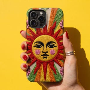 May include: A phone case featuring an embroidered sun design with a human face, pink cheeks, and radiating beams in shades of red, orange, yellow, and green. The background of the embroidery is a cream-colored knit texture with small pink flowers.