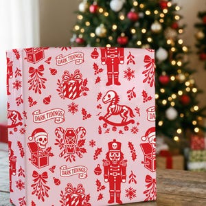 Premium satin finish wrapping paper featuring a repeating pastel goth pattern on a pink background. The design showcases a playful yet spooky aesthetic with high-contrast skeleton nutcrackers, skull jack-in-the-boxes, and rocking horse skeletons in soft pinks, and reds. The paper texture appears semi-glossy. Shown in three available sizes including sheets and rolls, specifically 29 inches wide, suitable for wrapping gifts for holidays, birthdays, or gothic themed occasions.