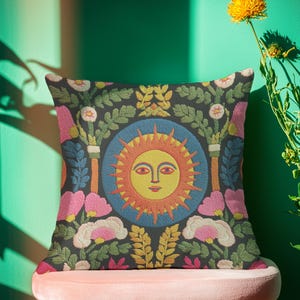 A square folk art sun throw pillow with a dark, charcoal-gray background. The central design is a vibrant celestial sun face in shades of orange, yellow, and red, with defined facial features. The sun is enclosed in a circular blue border with a textured, faux-embroidery pattern. Surrounding the central sun are symmetrical, colorful botanical and floral elements, including pink and white flowers, and green and gold leaves. The overall style is maximalist and bohemian.