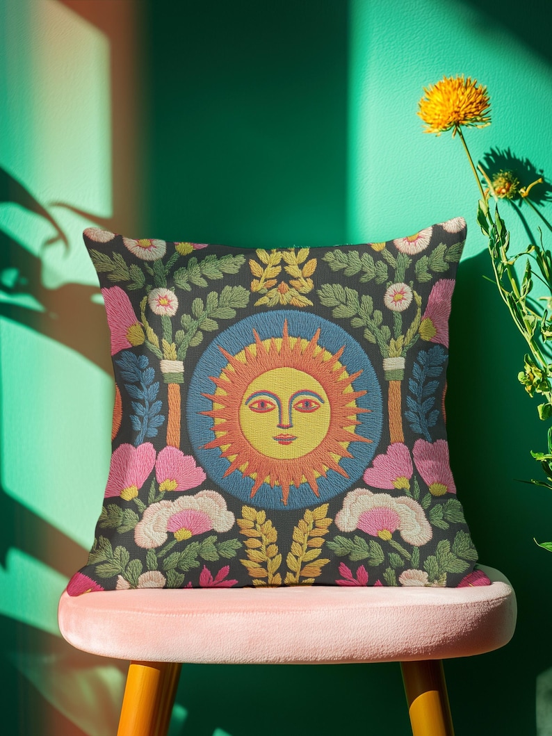 A square folk art sun throw pillow with a dark, charcoal-gray background. The central design is a vibrant celestial sun face in shades of orange, yellow, and red, with defined facial features. The sun is enclosed in a circular blue border with a textured, faux-embroidery pattern. Surrounding the central sun are symmetrical, colorful botanical and floral elements, including pink and white flowers, and green and gold leaves. The overall style is maximalist and bohemian.