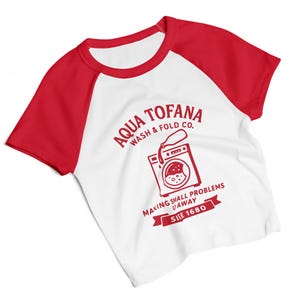 A women's fitted, mid-length micro rib raglan baby tee. The tee's body is solid white, made from a 1x1 micro-rib fabric.  It features short, set-in sleeves and a crewneck collar binding in a bold, contrasting athletic red. Centered on the chest is a vintage-inspired graphic printed in the same red ink. The design is for the "Aqua Tofana Wash & Fold Co." and features an illustration of an old-fashioned washing machine. Text below the machine reads "Making small problems go away" and "Since 1680."