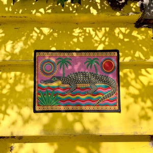 A rectangular doormat featuring a colorful 2D flat print designed to mimic raised, textured yarn embroidery. The central graphic showcases a large dark grey and yellow alligator resting in wavy water stripes of red, bright blue, and yellow. The background is hot pink, decorated with two green palm trees, a leafy green agave plant, and two stylized suns. The entire design is enclosed by a yellow border with repeating circle motifs and finished with a sleek black outer edge border trim.