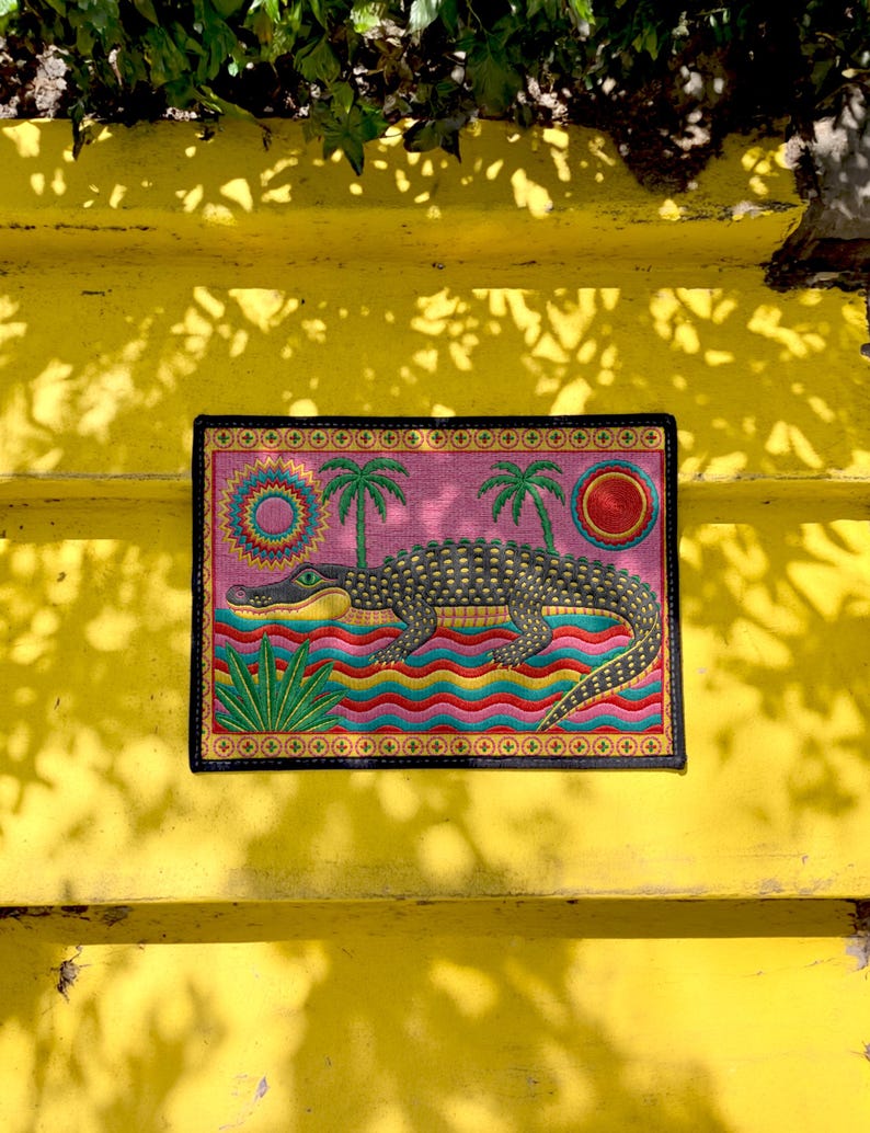 A rectangular doormat featuring a colorful 2D flat print designed to mimic raised, textured yarn embroidery. The central graphic showcases a large dark grey and yellow alligator resting in wavy water stripes of red, bright blue, and yellow. The background is hot pink, decorated with two green palm trees, a leafy green agave plant, and two stylized suns. The entire design is enclosed by a yellow border with repeating circle motifs and finished with a sleek black outer edge border trim.