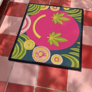 A rectangular doormat featuring a large hot pink smiley face on a deep teal background. The smiley face has green cannabis leaves for eyes and a yellow stitched-style smile. The central face is surrounded by stylized green foliage arches and concentric circles in pink, yellow, and white, resembling abstract flowers. The design mimics a vintage needlepoint or tapestry texture but is a flat print. The mat has a solid black trim border.