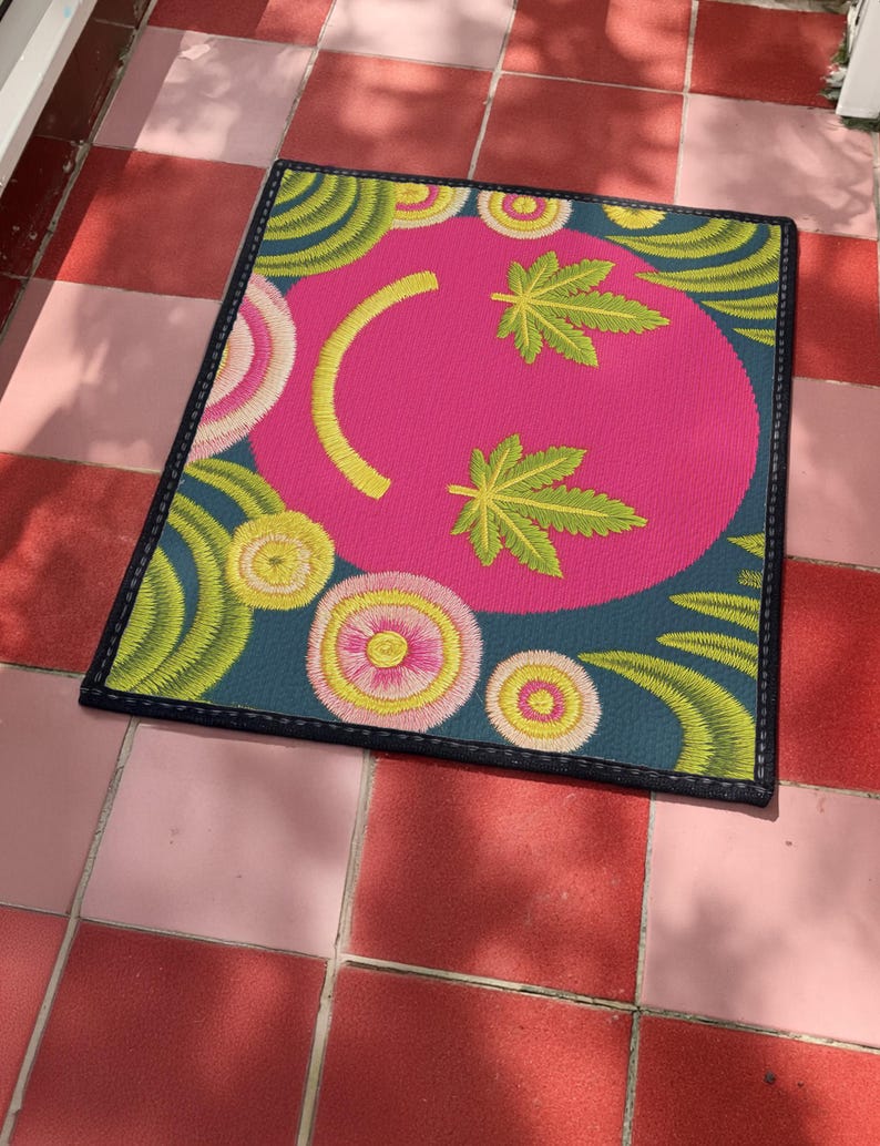 A rectangular doormat featuring a large hot pink smiley face on a deep teal background. The smiley face has green cannabis leaves for eyes and a yellow stitched-style smile. The central face is surrounded by stylized green foliage arches and concentric circles in pink, yellow, and white, resembling abstract flowers. The design mimics a vintage needlepoint or tapestry texture but is a flat print. The mat has a solid black trim border.