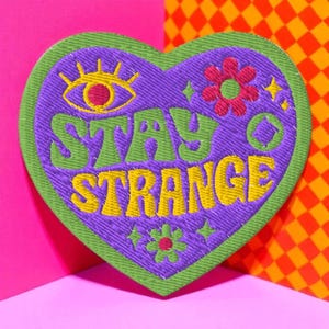May include: Embroidered heart-shaped patch with the words "STAY STRANGE" in yellow and green. The patch features a purple background with a green border, an eye, flowers, and stars.