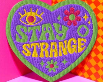 Könnte beinhalten: Gestickter herzförmiger Aufnäher mit den Worten "STAY STRANGE" in Gelb und Grün. Der Aufnäher hat einen lila Hintergrund mit einem grünen Rand, ein Auge, Blumen und Sterne.